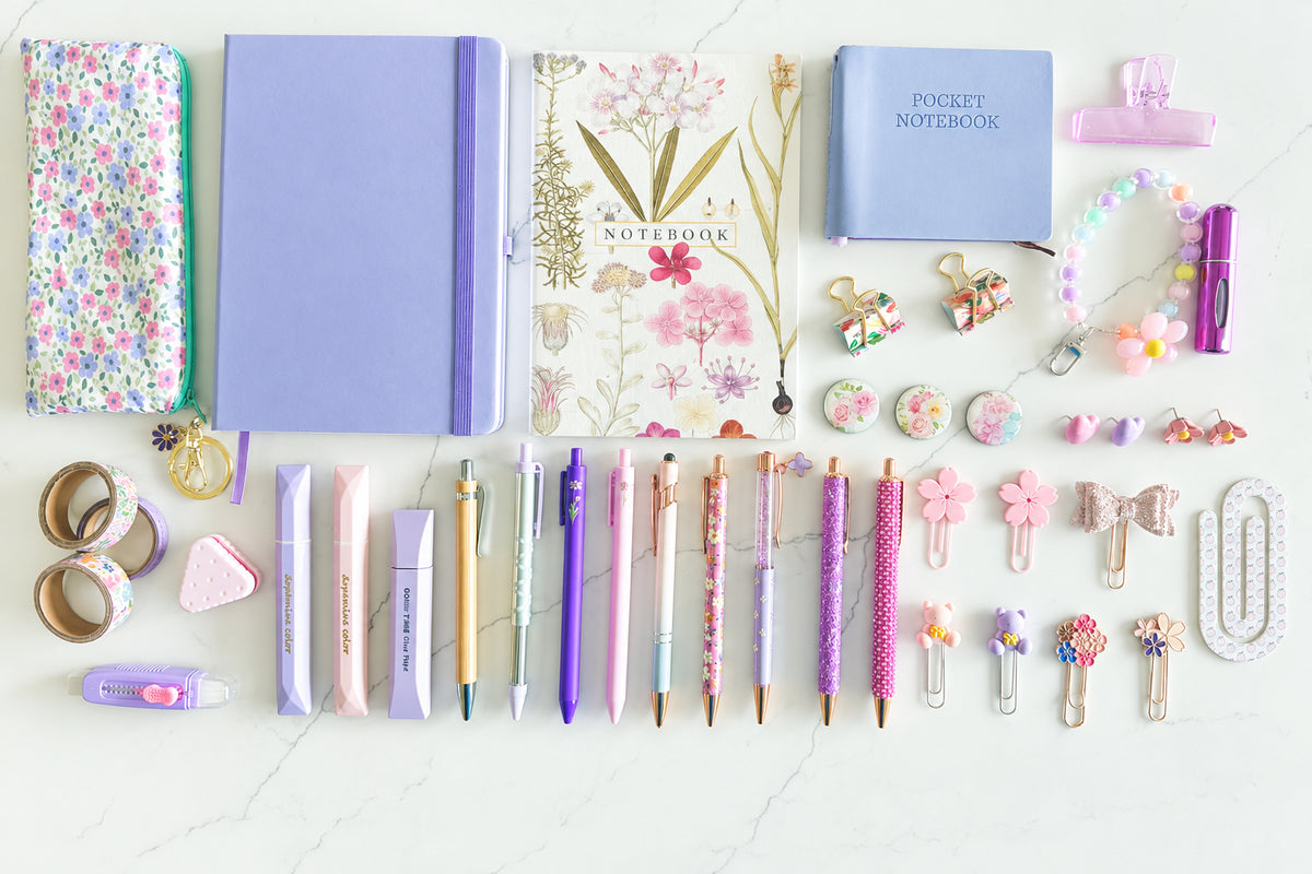 A flat-lay collection of pastel stationery and desk accessories arranged on a light surface, including notebooks, pens, clips, washi tape, buttons, and small decorative floral items in pink, purple, blue, and cream tones.